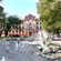 Mature trees and a large fountains with bronze statues in front of a monumental building